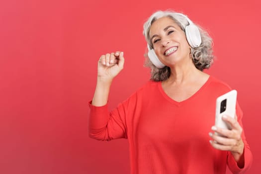 Studio portrait with red background of a happy mature woman dancing and listening to music with a mobile