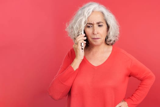 Studio portrait with red background of an angry mature woman talking on the mobile phone
