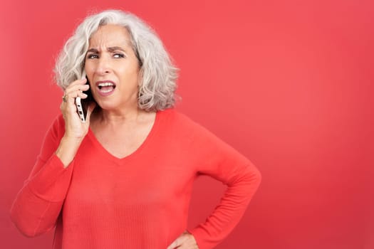 Studio portrait with red background of a mature woman trying to talk on her mobile phone but she can't hear anything