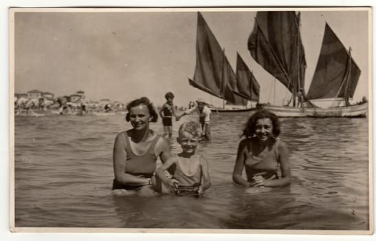 ITALY - CIRCA 1940s: Vintage photo shows family - mother with son and daughter) have a swimm in the sea. Sailing boats are on the background. Vacation theme. Retro black and white photography.