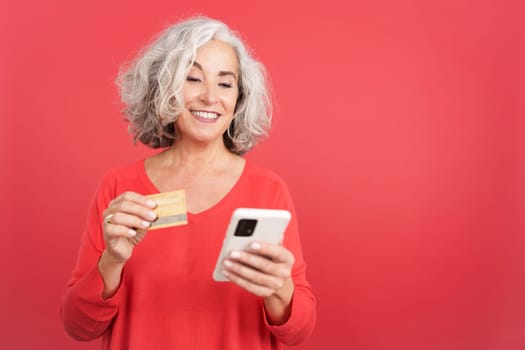Studio portrait with red background of a mature woman using a credit card to shopping online with the mobile