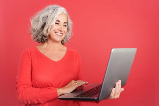 Studio portrait with red background of a mature woman smiling and using a laptop
