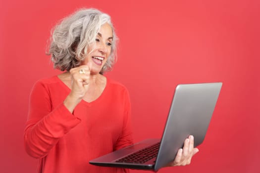 Studio portrait with red background of a mature woman celebrating while using a laptop