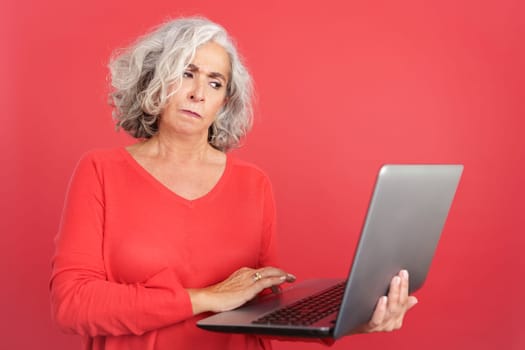 Studio portrait with red background of a worried mature woman standing and using a laptop