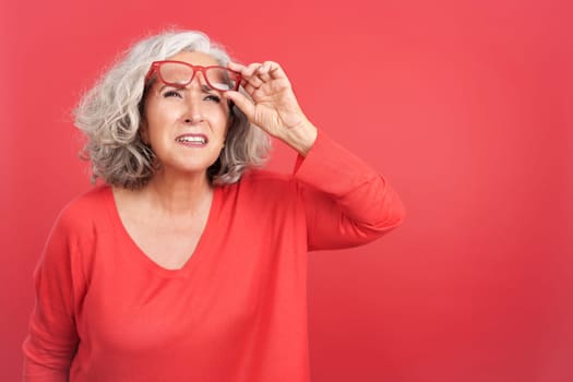 Studio portrait with red background of a mature woman lifting up her glasses to see from a distance