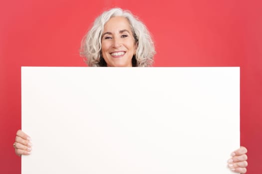 Studio portrait with red background of a mature smiley woman holding a blank panel. Mock up