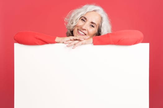 Studio portrait with red background of a relaxed mature woman leaning on a blank panel