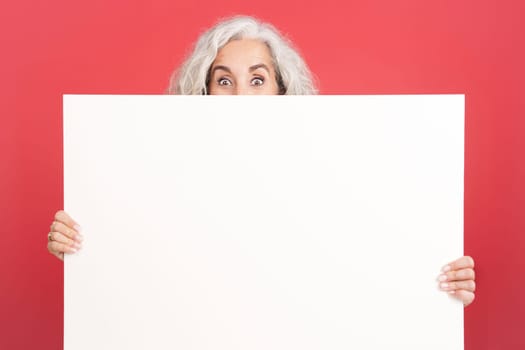 Studio portrait with red background of a mature woman hiding behind a blank panel