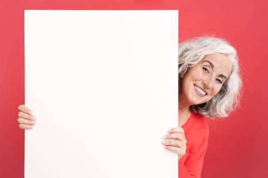 Studio portrait with red background of a smiley mature woman holding a blank panel
