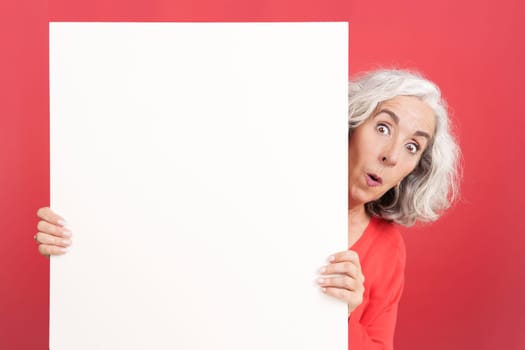Studio portrait with red background of a surprised mature woman holding a blank board