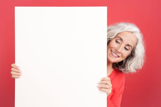 Studio portrait with red background of a mature woman looking at the white board that she is holding