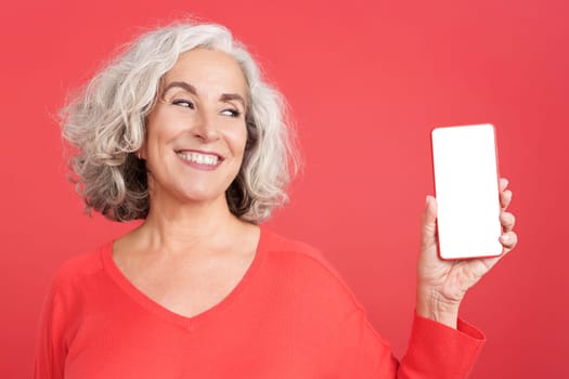 Studio portrait with red background of a mature woman showing the blank screen of the mobile. Mock up