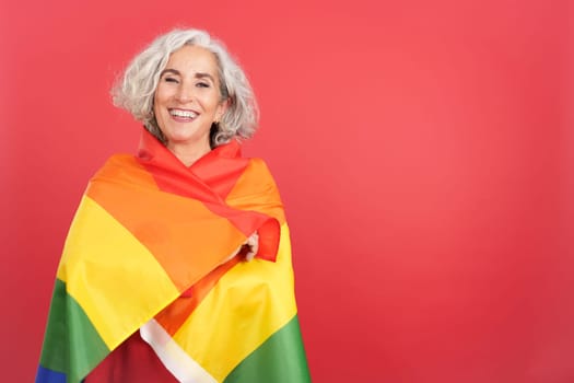 Studio portrait with red background of a mature woman wrapped with a lgbt rainbow flag