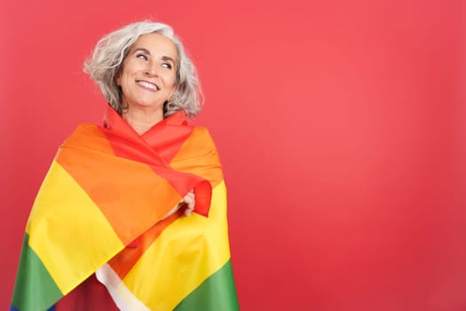 Studio portrait with red background of a smiley mature woman wrapped with a lgbt rainbow flag
