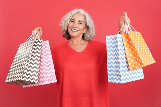 Studio portrait with red background of a happy mature woman standing showing shopping bags