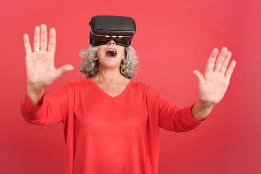 Studio portrait with red background of a surprised mature woman using virtual reality goggles