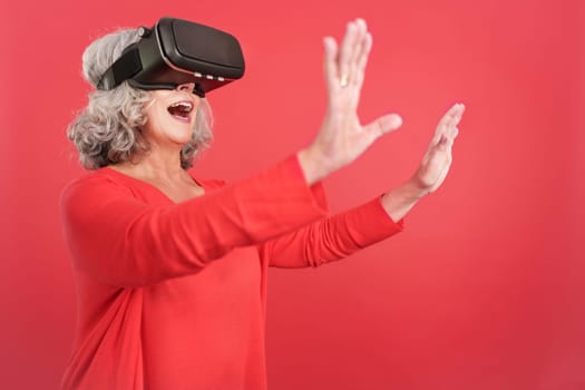 Studio portrait with red background of a mature woman gesturing while using a virtual reality goggles
