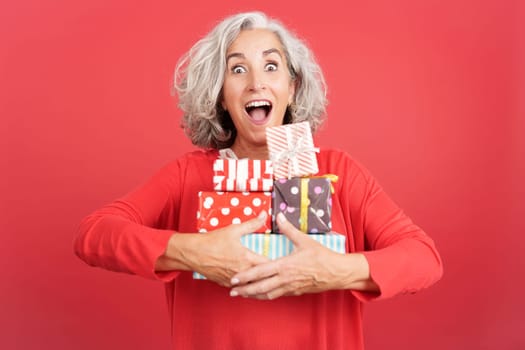 Studio portrait with red background of an excited mature woman receiving many gifts