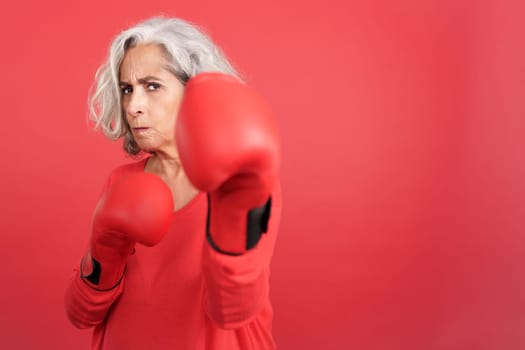 Studio portrait with red background of a mature woman in fighting pose wearing boxing gloves