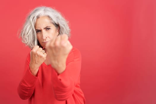 Studio portrait with red background of a mature woman confronting the camera with her fists
