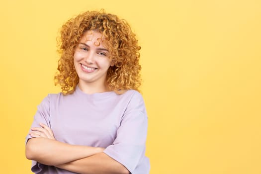 Studio image with yellow background of a smiley woman with curly hair looking at camera standing with arms crossed