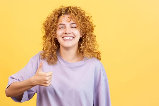 Studio image with yellow background of a happy young woman with curly hair gesturing agreement raising the thumb