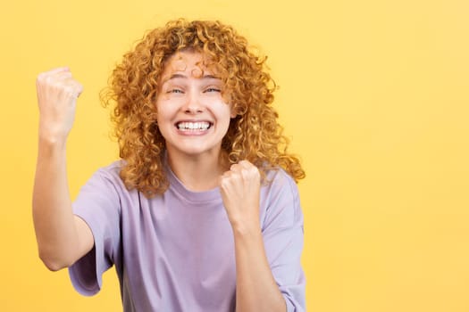 Studio image with yellow background of a beauty woman with curly hair looking at camera while celebrating raising the fists