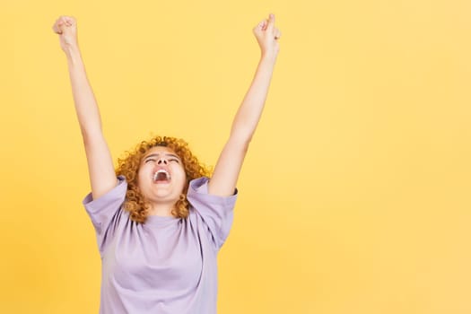 Studio image with yellow background of a happy woman with curly hair celebrating while raising the arms and shouting