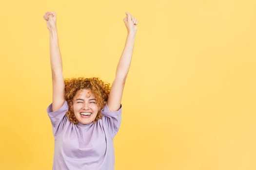 Studio image with yellow background of a happy woman with curly hair celebrating while raising the arms and smiling