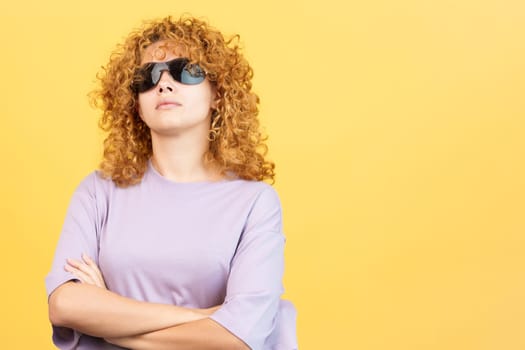 Studio image with yellow background of a rude woman with curly hair and sunglasses standing with arms crossed