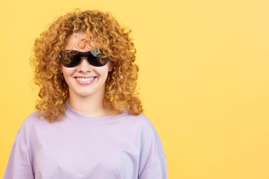 Studio image with yellow background of a happy woman with curly hair and sunglasses looking at the camera