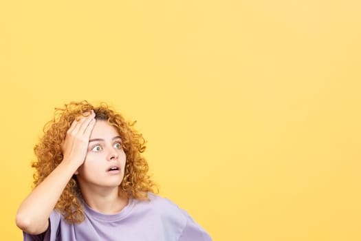 Studio image with yellow background of a woman with curly hair gesturing with the hand on head expressing despair