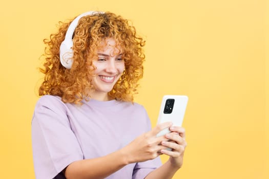 Studio image with yellow background of a woman with curly hair listening to music smiling with a mobile and headphones