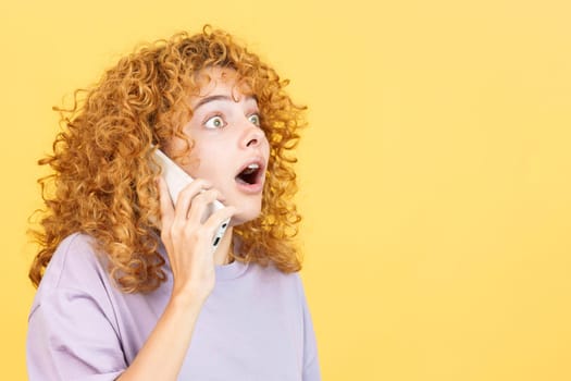 Studio image with yellow background of a surprised young woman with curly hair open the mouth while talking to the mobile