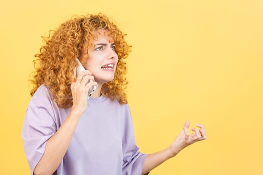 Studio image with yellow background of an angry woman with curly hair talk to the mobile phone