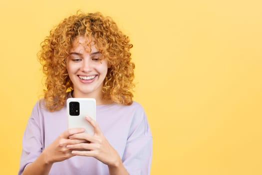 Studio image with yellow background of a beauty and happy woman with curly hair using the mobile phone