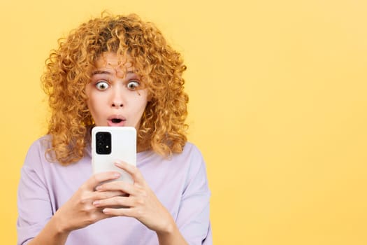 Studio image with yellow background of a surprised woman with curly hair looking the screen of the mobile phone