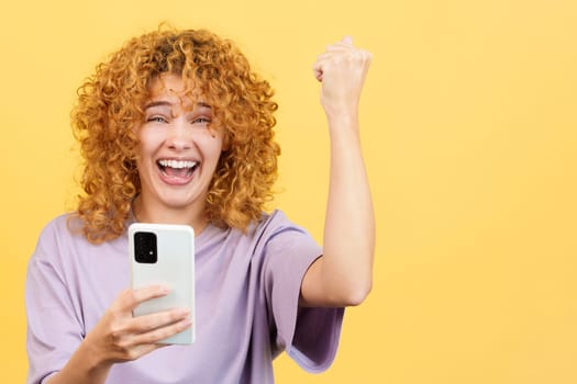 Studio image with yellow background of an excited woman with curly hair celebrating while using the mobile phone