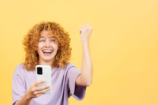 Studio image with yellow background of a happy woman with curly hair celebrating while using the mobile phone