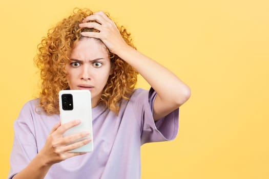 Studio image with yellow background of a desperate woman with curly hair looking at mobile phone and gesturing