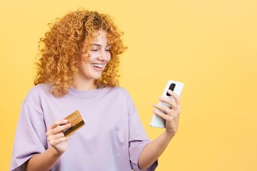 Studio image with yellow background of a smiley woman with curly hair using a card to shopping online with the mobile