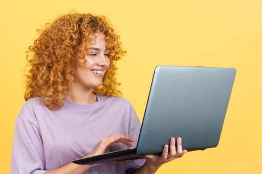 Studio image with yellow background of a beauty woman smiling with curly hair standing and using a laptop