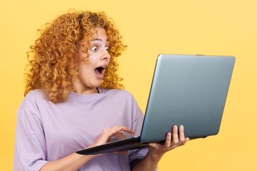 Studio image with yellow background of a beauty surprised woman with curly hair using a laptop while standing