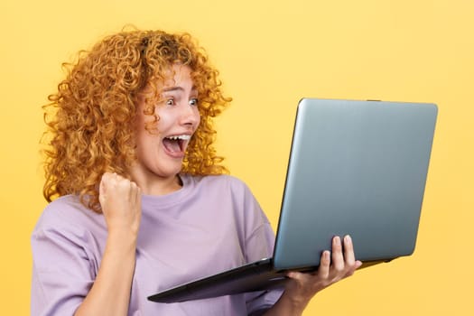 Studio image with yellow background of a emotional happy woman with curly hair celebrating while using a laptop