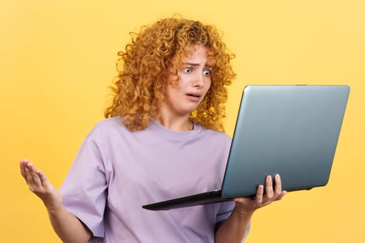 Studio image with yellow background of a worried woman with curly hair standing while holding a laptop expressing a problem