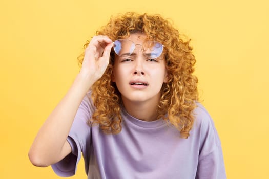 Studio image with yellow background of a young woman taking off her glasses to see far away while looking at camera