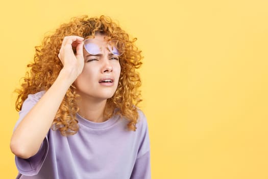 Studio image with yellow background of a woman with curly hair taking off her glasses to see far away but finds it hard to do so