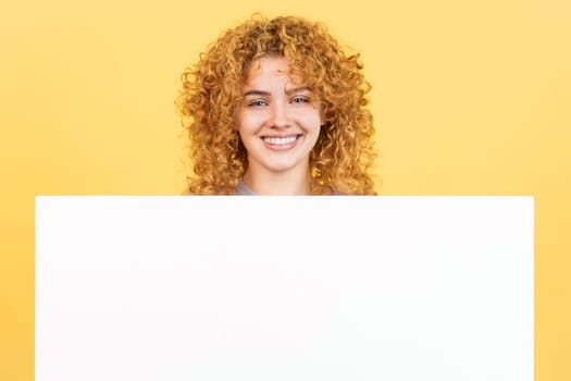 Studio image with yellow background of a beauty and smiley woman with curly hair holding a blank panel
