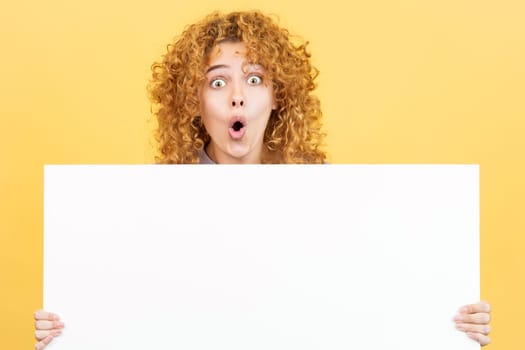Studio image with yellow background of a woman with curly hair holding a blank panel while expressing surprise