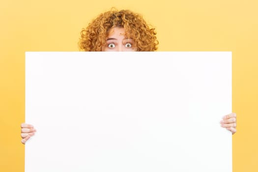 Studio image with yellow background of a surprised woman with curly hair hiding behind a blank panel